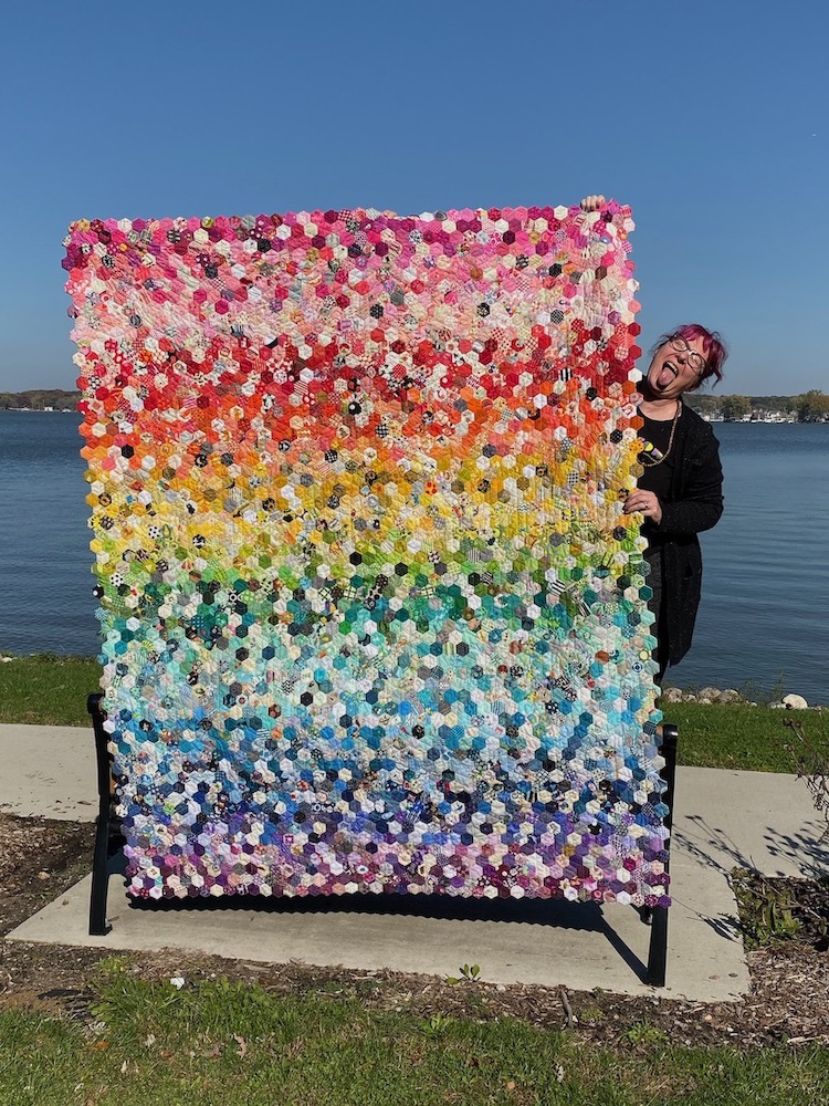 a white women with pink hair and glasses sticks out her tongue as she stands next to a quilt made up of a rainbow of 1-inch hexagons