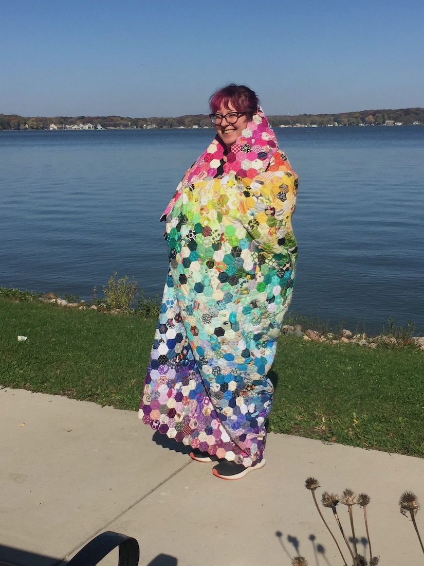 a white woman with pink hair and glasses stands next to a lake, draped in a rainbow hexie quilt