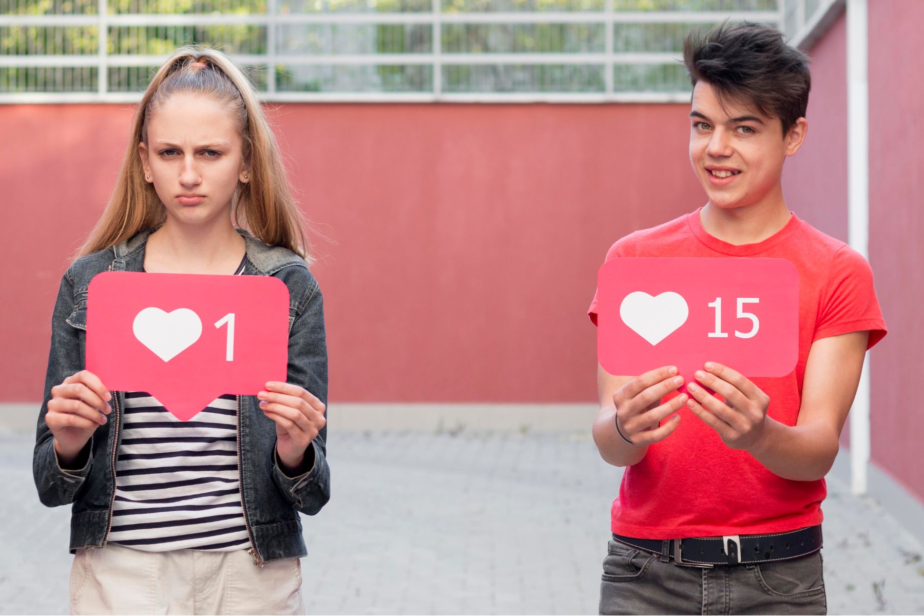 A young white woman with blond hair holds a pretend Instagram “like count” of 1 while a young white person with dark, short hair holds up a pretend Instagram “like count” of 15