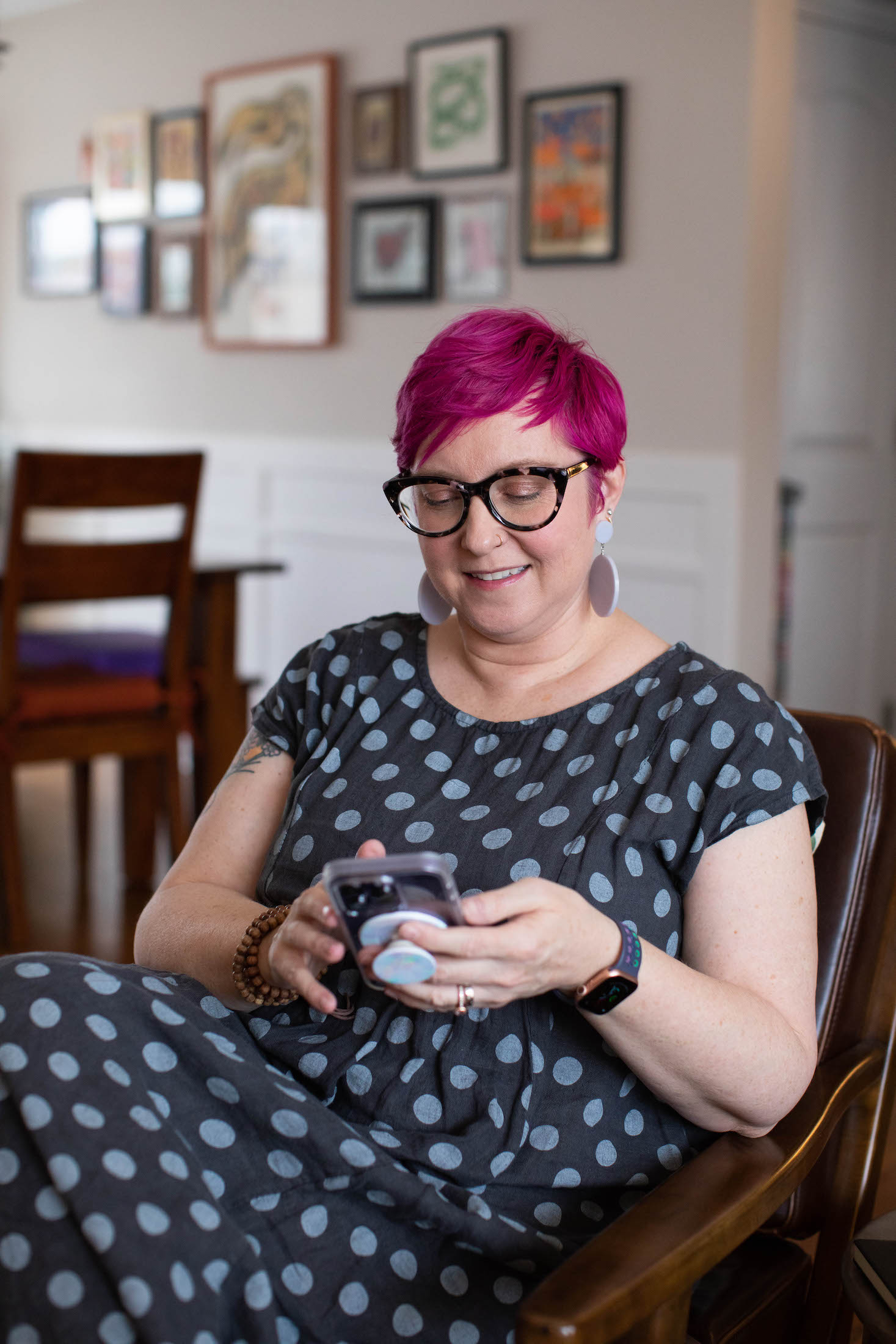 White woman with pink pixie cut and glasses sits on a chair in her living room, looking at her phone