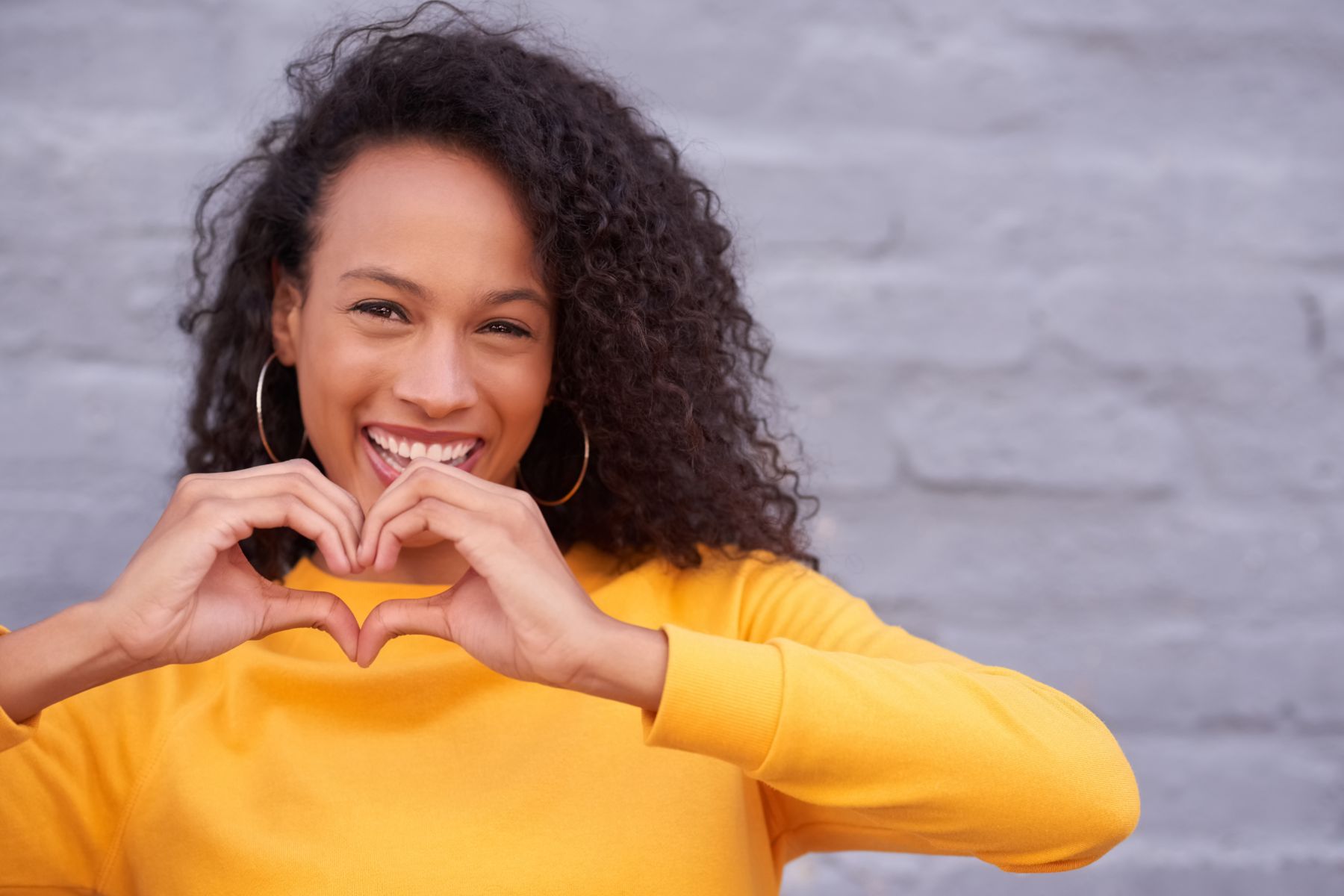 Una joven negra con el pelo rizado hasta los hombros y una camiseta amarilla levanta las manos en forma de corazón